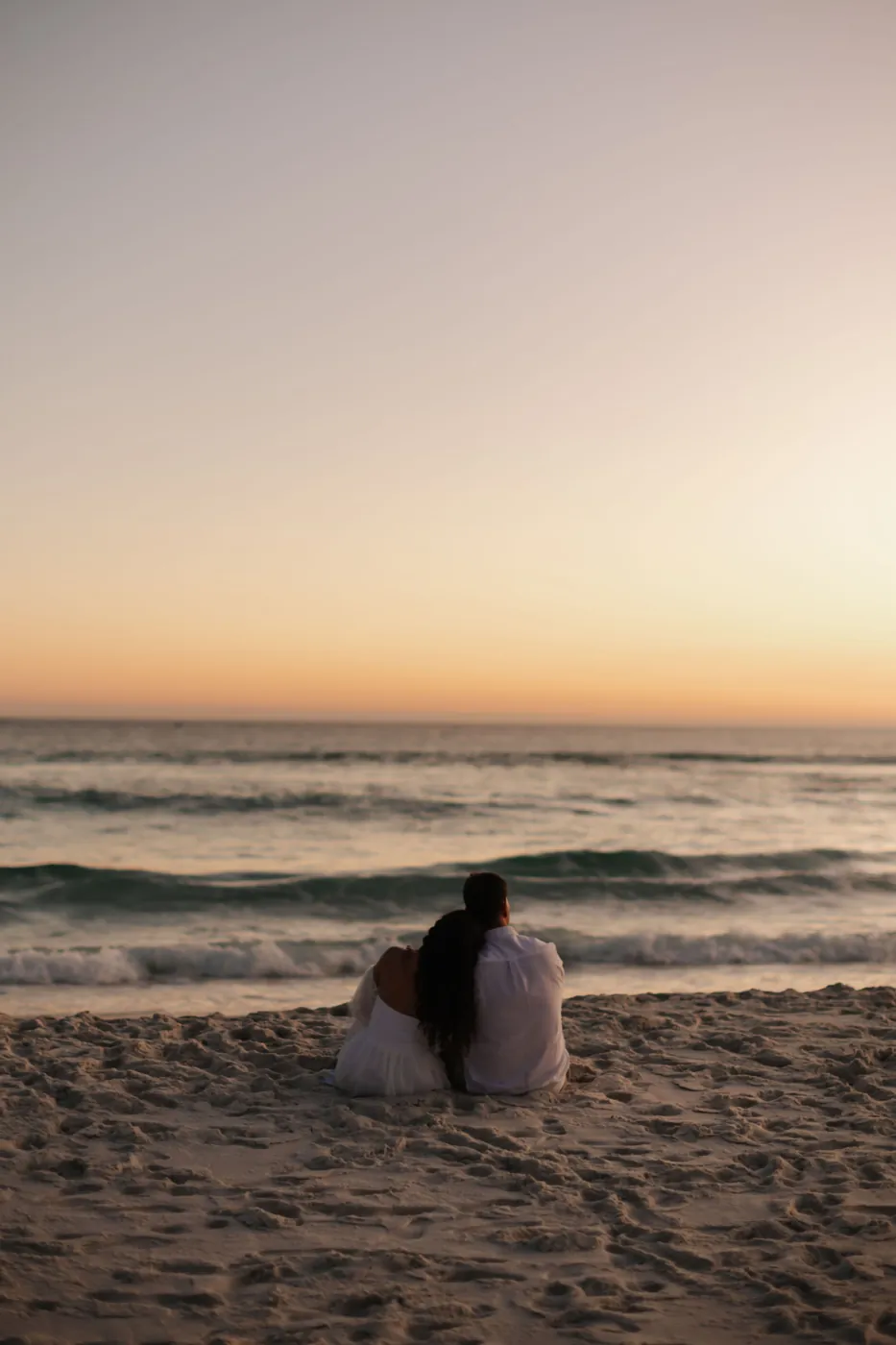 Casal sentado na areia, abraçado, olhando o mar