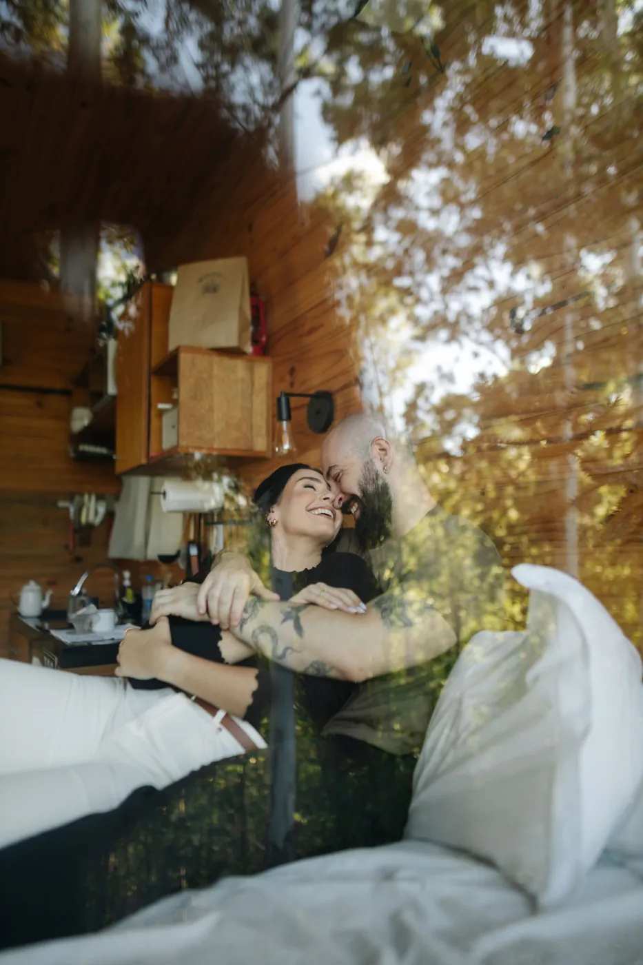 Casal rindo abraçado, visto pelo reflexo da janela da cabana