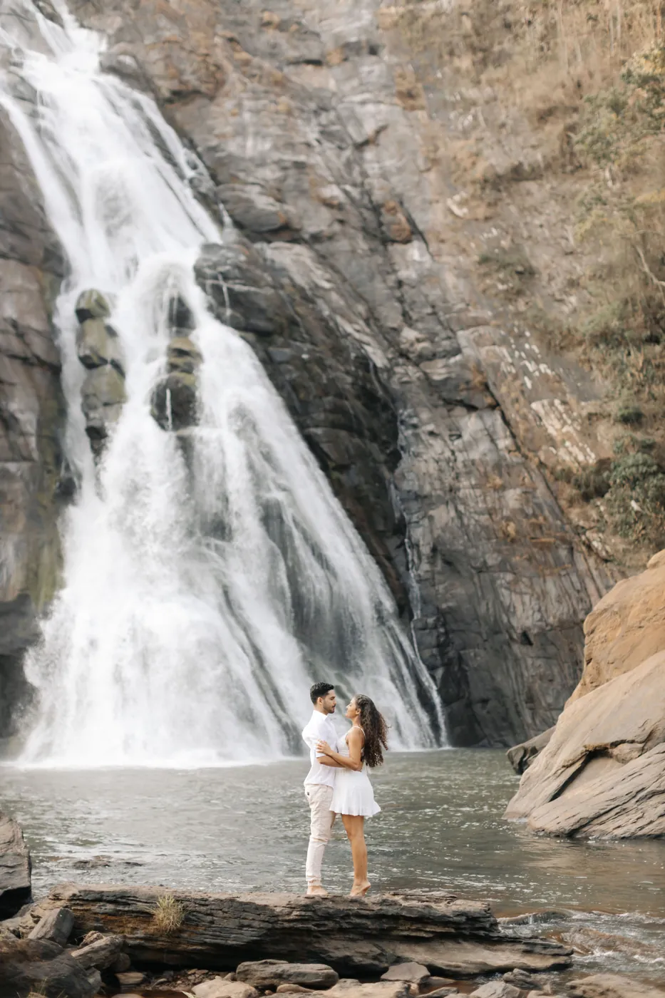 Casal abraçado sobre uma pedra na frente da queda d’água
