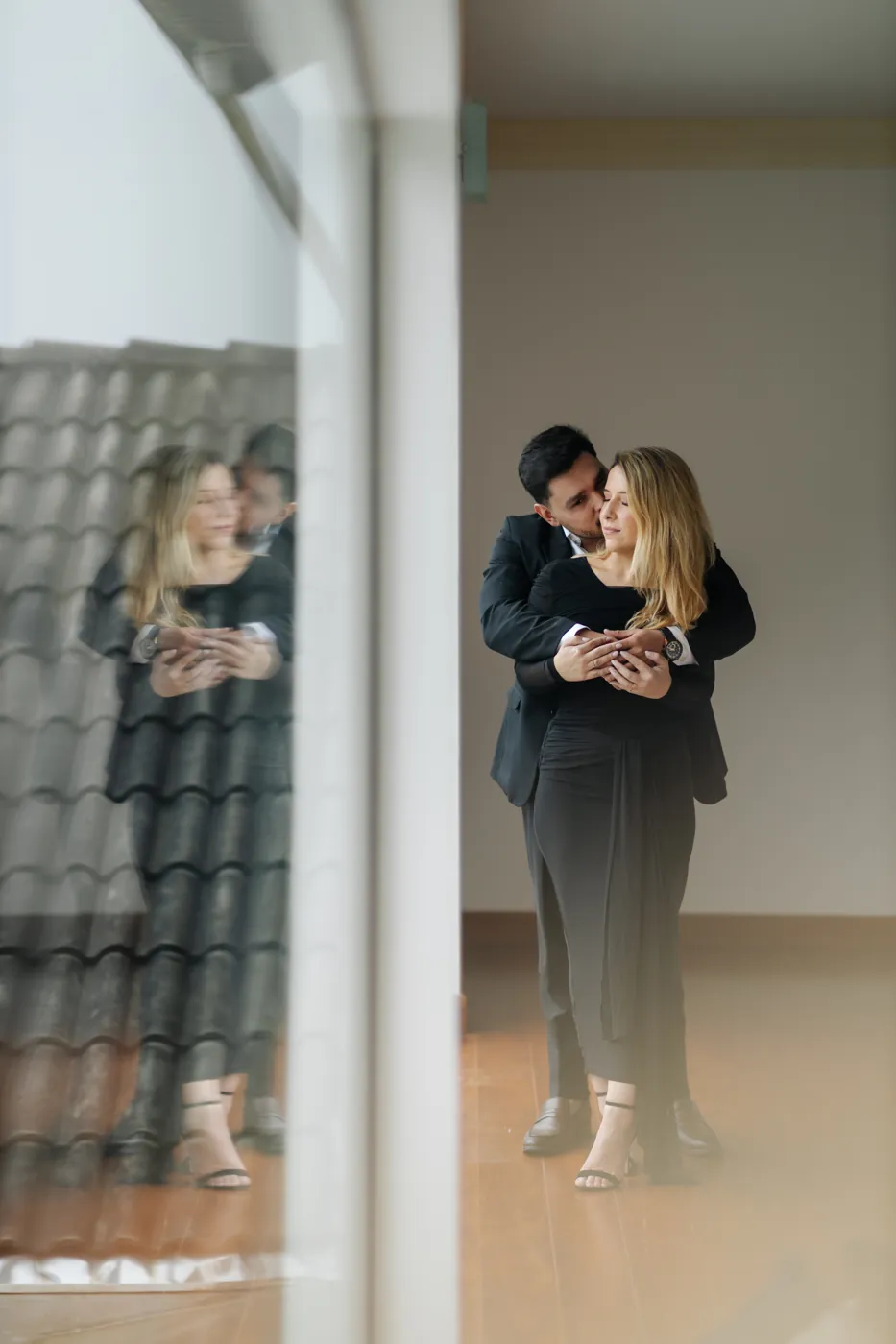 Ele beija o rosto dela, os dois refletidos no vidro da varanda
