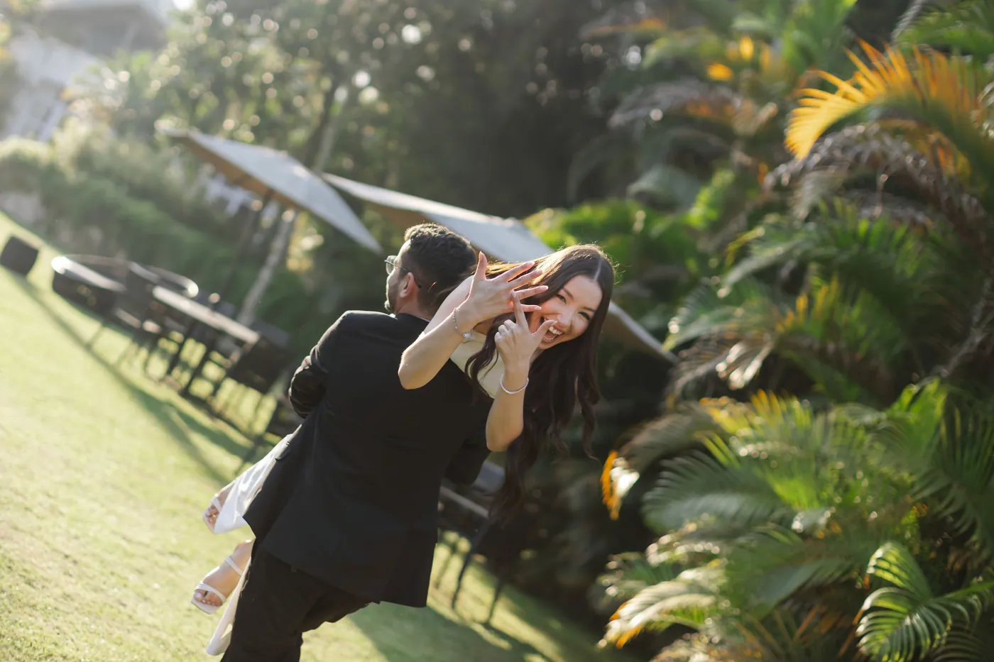 Ela mostrando o anel, rindo, enquanto ele a levanta no jardim