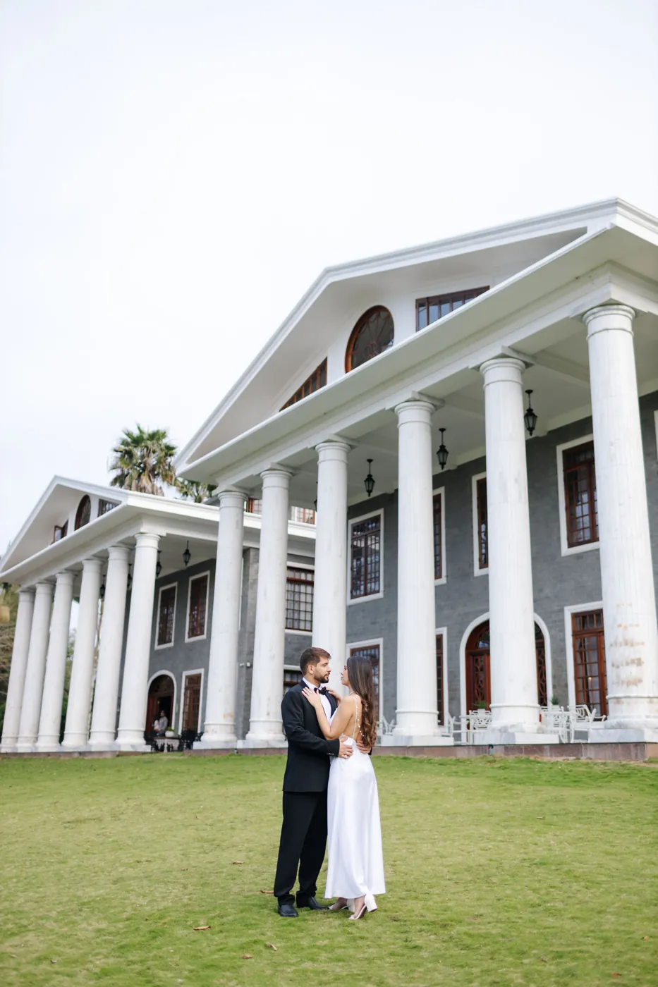 Casal abraçado no gramado em frente às colunas brancas do hotel