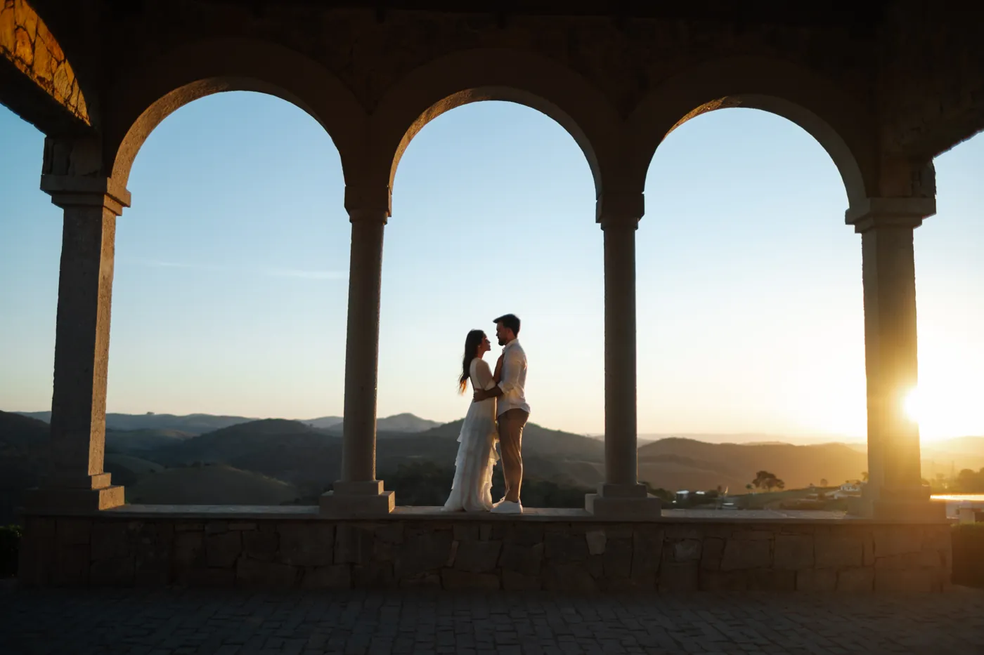 Casal abraçado sob os arcos de pedra, com as montanhas no pôr do sol