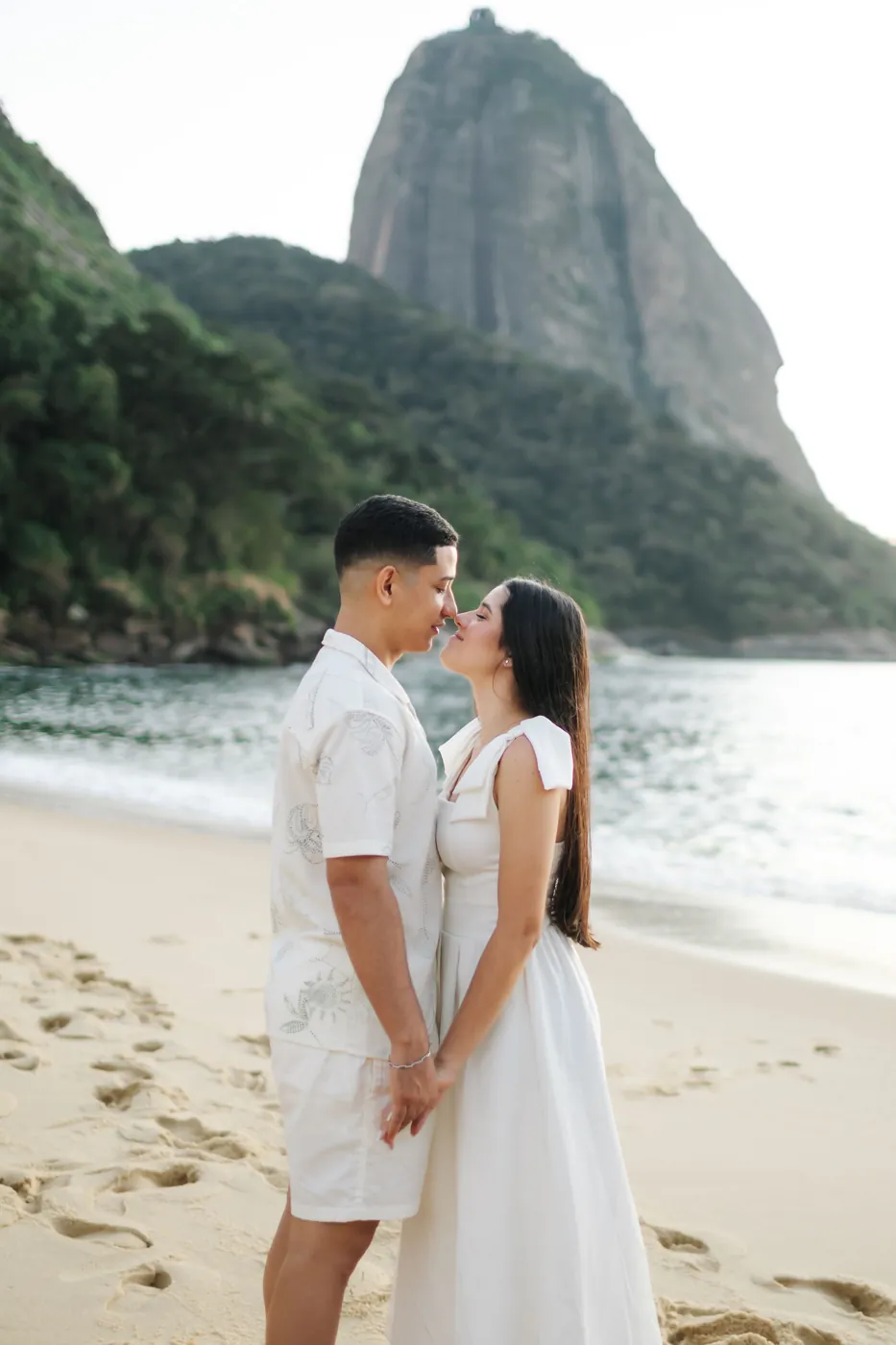 Casal quase se beijando na areia com o Pão de Açúcar ao fundo