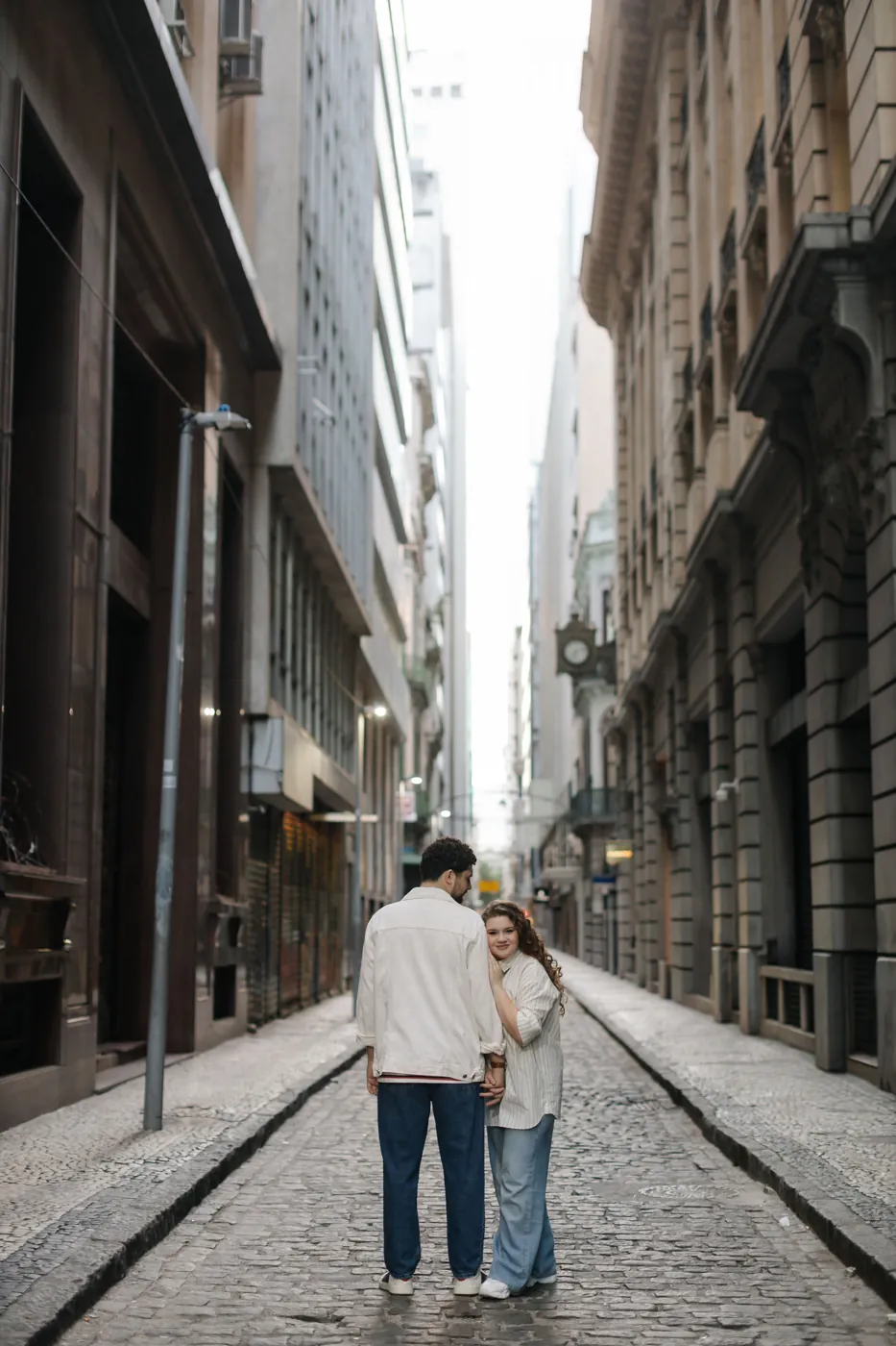 Casal de costas numa rua estreita, ela olhando para a câmera