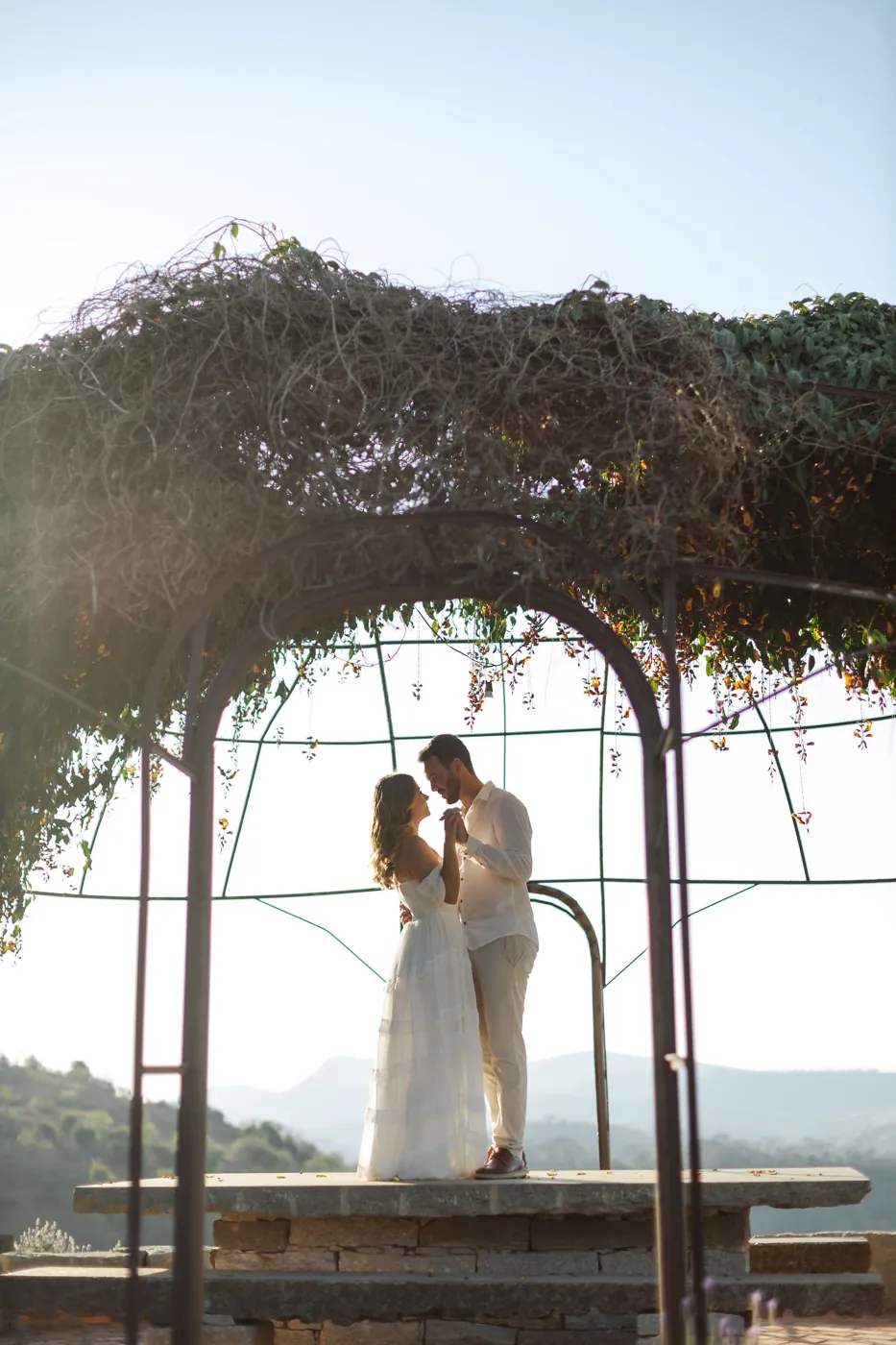 Casal dançando sob um gazebo coberto de trepadeira, com as montanhas ao fundo