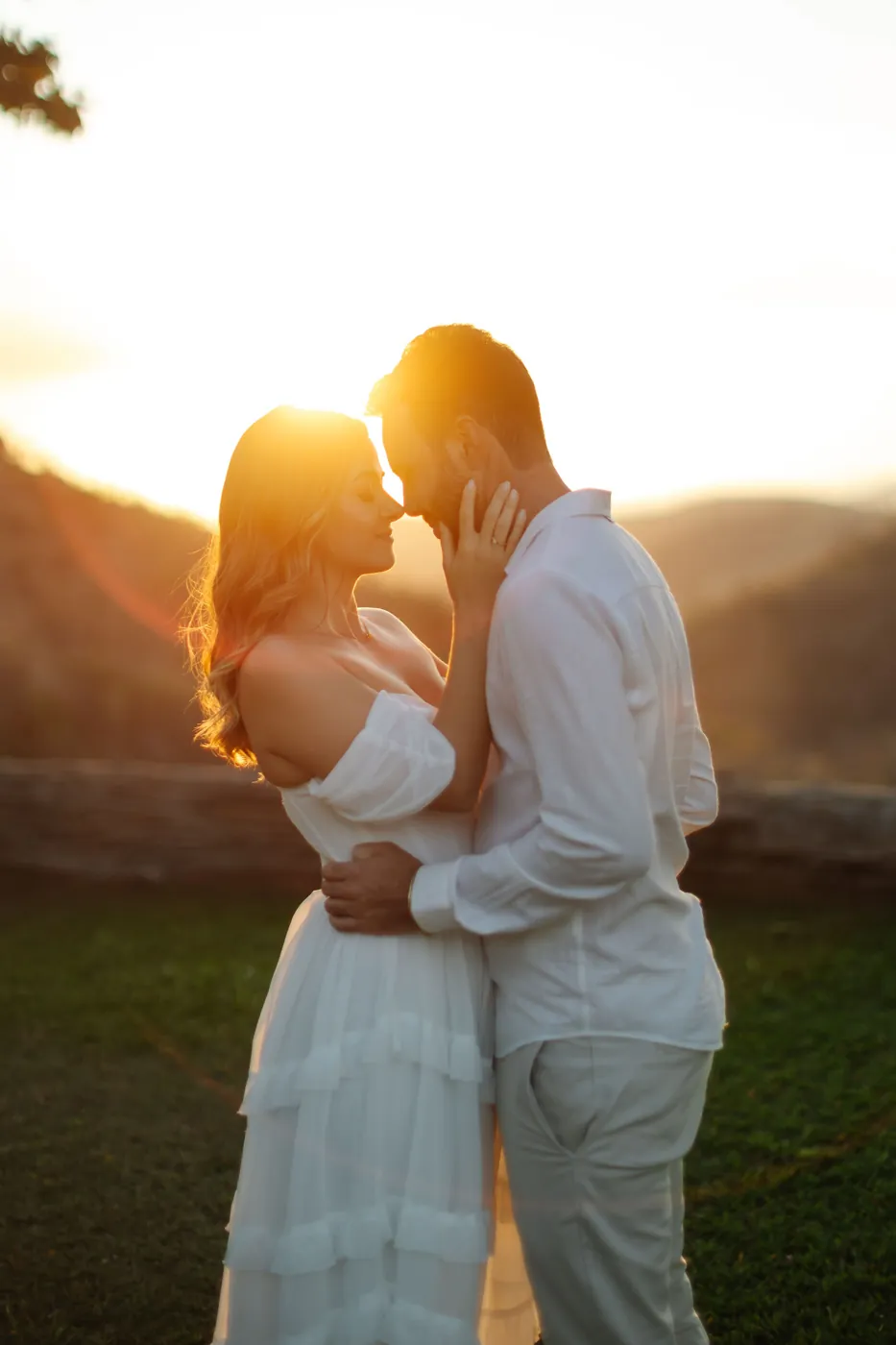 Casal quase se beijando, ela com a mão no rosto dele, no pôr do sol